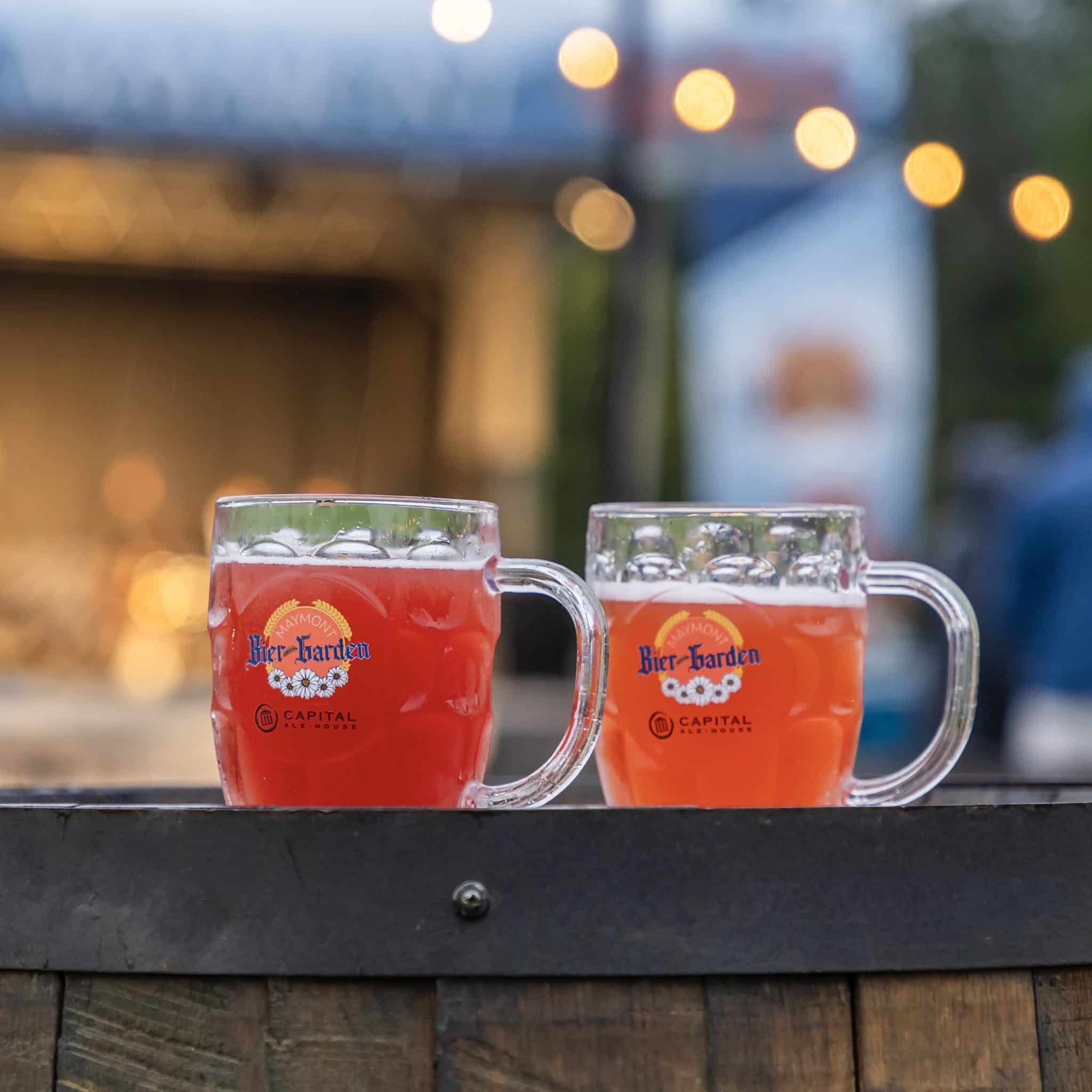 Two glass mugs with the Bier-Garden logo are full of a reddish beer, sitting on a barrel with a stage and lights in the background